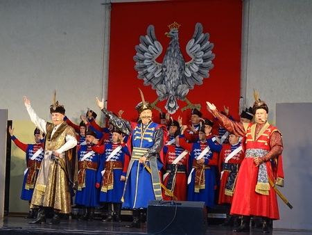 na scenie amfiteatru w Hajnówce stoi grupa artystów w strojach historycznych. w tle widać duże godło Polski