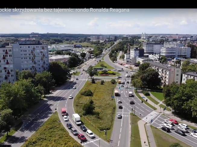 w środkowej części przebiegają ulice z licznymi samochodami. Z lewej strony wysokie bloki. Na zdjęciu widać dużo zieleni miejskiej.