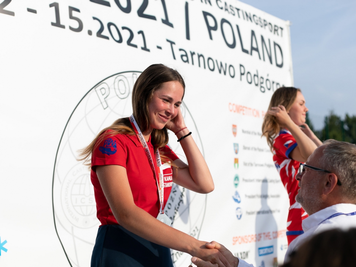 młoda dziewczyna w czerwonej koszulce stoi na podium; jej rękę ściska mężczyna; za nia biała plansza promocyjna