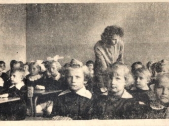 Dzieci siedzą w klasie przy stolikach, pomiędzy nimi nauczycielka