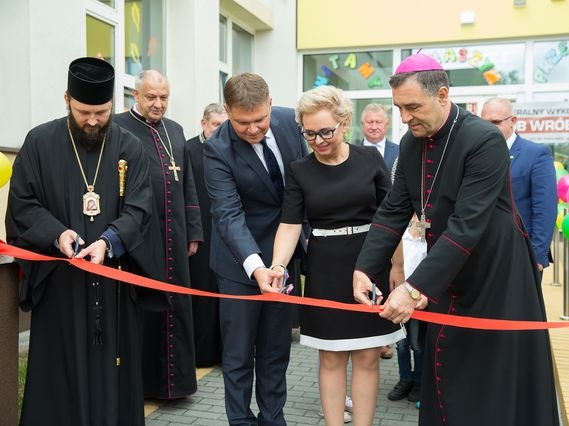 Zdjęcie przedstawia moment uroczystego przecięcia wstęgi przez panią dyrektor przedszkola, przedstawicieli władz miasta, duchownych obu wyznań przed wejściem do nowej części przedszkola. 