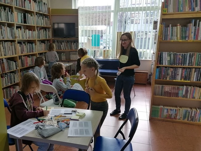 Grupa dzieci siedzi przy stolikach. W tle widać książki stojące na półkach.