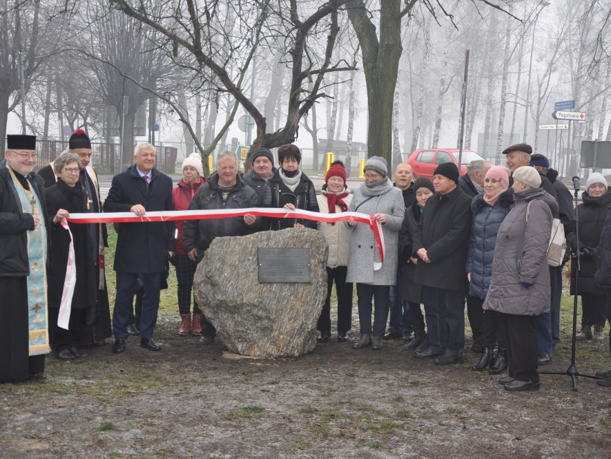 Zdjęcie grupowe, uczestnicy uroczystości stoją po obu stronach kamienia z tablicą, trzymając biało - czerwoną wstęgę