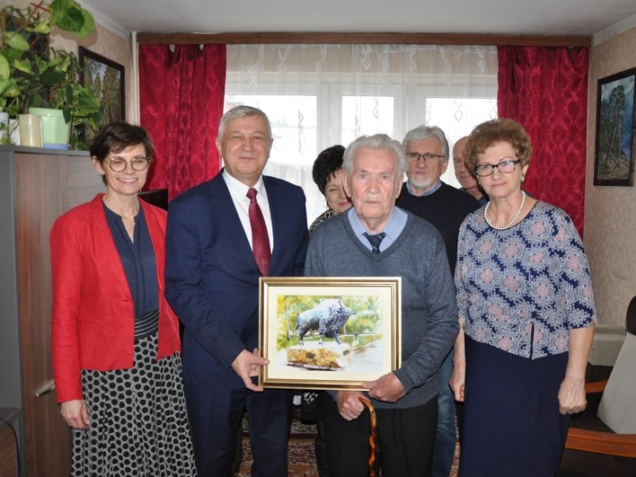 Jubilat z burmistrzem trzymają wsólnie obraz przedstawiający żubra. Obok stoi rodzina i kierownik Urzędu Stanu Cywilnego w Hajnówce