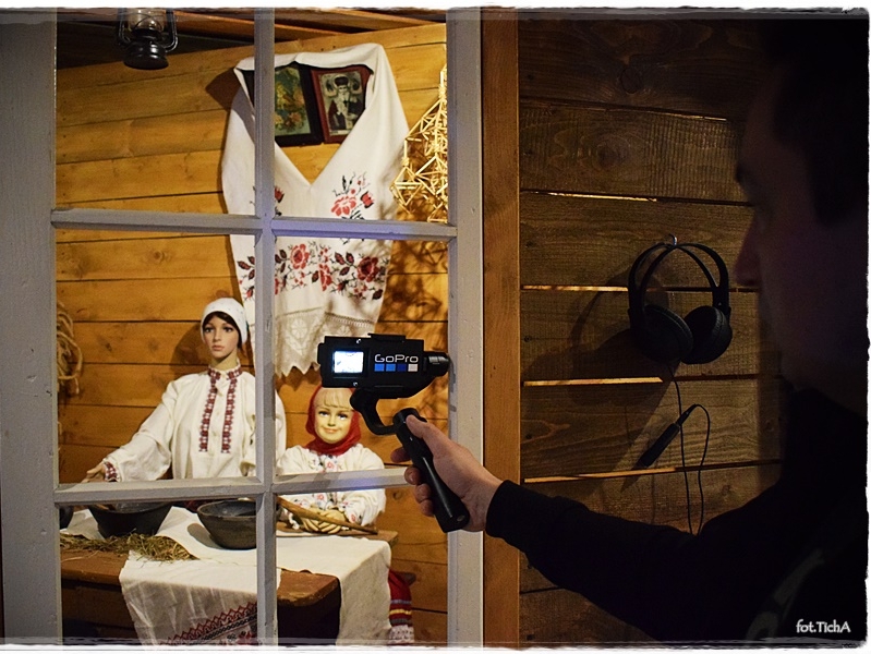 Z prawej strony stoi osoba trzymająca kamerę nakierowana na wystawę manekinów w scenerii białoruskiej chaty.