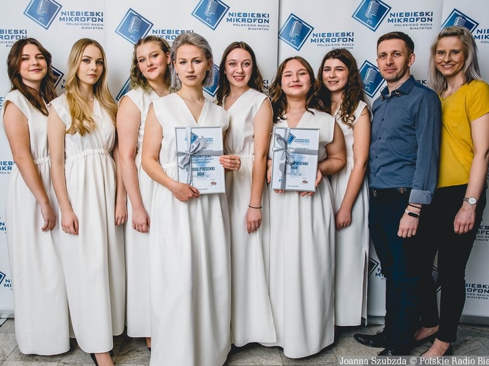 Dziewczęta ze Studioa Piosenki HDK ubrane w białe długie suknie. Po prawej stronie stoi Przemysłam Zalewski i Marta Gredel - Iwaniuk