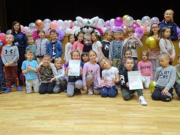 Duża grupa dzieci pozuje do zdjecia wraz z policjantkami i dużą żywą maskotką. W tle widać duzo balonów.