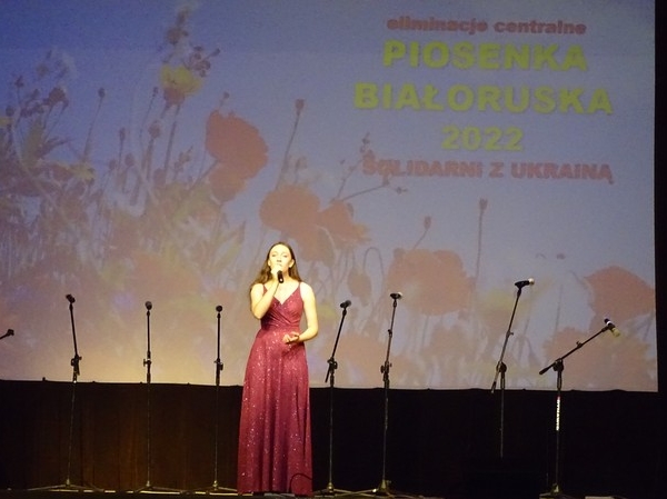 laureatka w czerwonej sukni śpiewa na scenie. W tle wyświetlony został duży slajd z tutułem konkursu