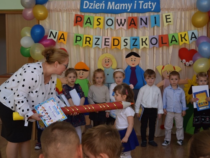 Pani dyrektor trzyma na ramieniu dziecka duży czerwony ołówek. Z tyłu stoi reszta grupy przedszkolnej.