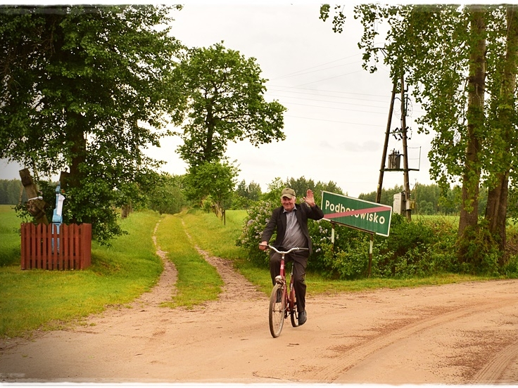 rowerzysta na drodze polnej. W tle widać znak drogowy z nazwą wsi.