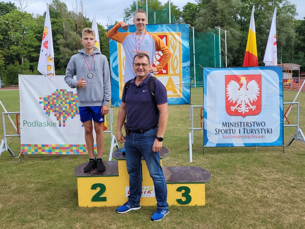 Zawodnicy na podium, przed stoi trener. W tle widać flagi, herby Ministerstwa Sportu i Turystyki oraz Województwa Podlaskiego 