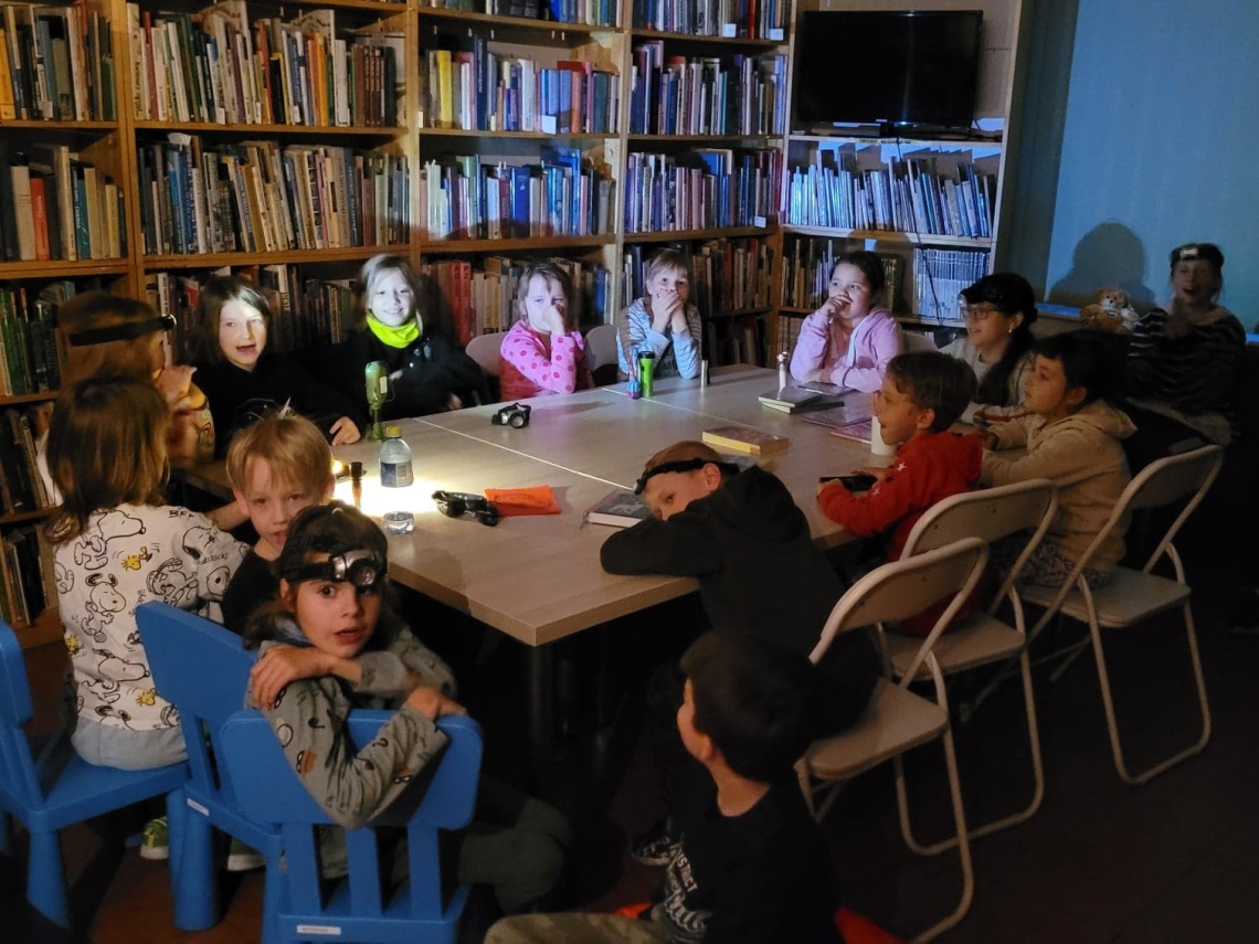dziei siedzą przy stole w ciemnej sali biblitecznej. Na głowach mają latarki czołowe.