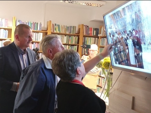 trzy osoby stoją przed telewizorem, na którym wyświewtlane jest stare zdjęcie
