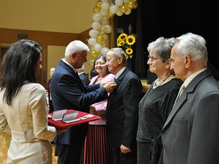 Burmistrz przypina mężczyźnie medal. Obok stoją Jubilaci. Młoda kobieta trzyma tacę z medalami.