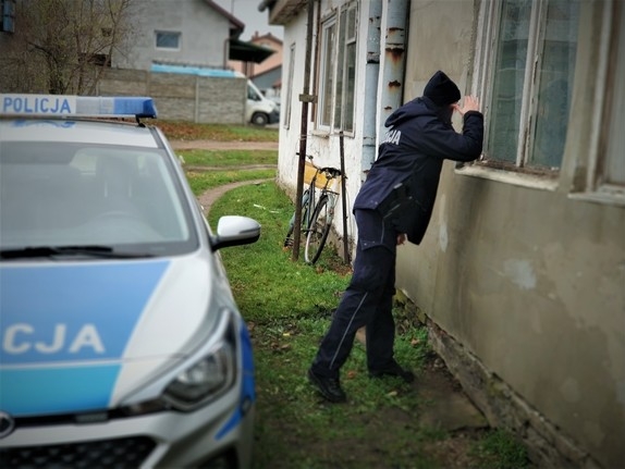 Policjant zagląd w okna budynku. Obok stoi zaparkowany radiowóz.