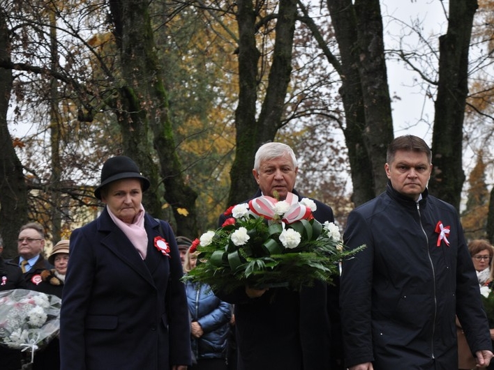 delegacja trzech osób .Męzczyzna w środku niesie wieniec z biało-czerwonych kwiatów. 