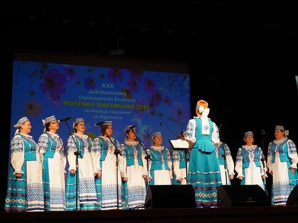 chór w białoruskich strojach stoi na scenie. W tle wyświetlany jest tytuł Festiwalu