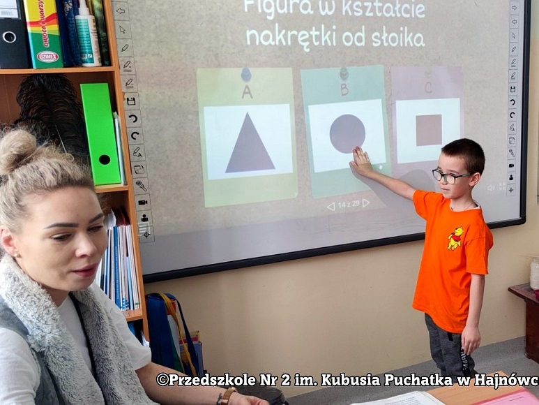 chłopiec pokazuje na tablicy figure w kształcie nagretki od słoika. Z lewej strony siedzi nauczyciel