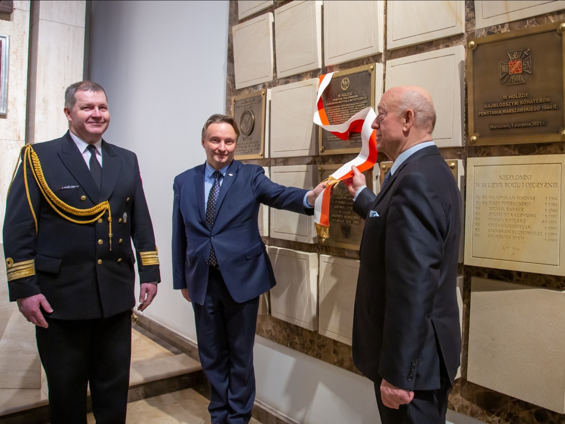 trzech mężczyzn dokonuje odsłonięcia tablicy pamiątkowej ulokowanej na ścianie