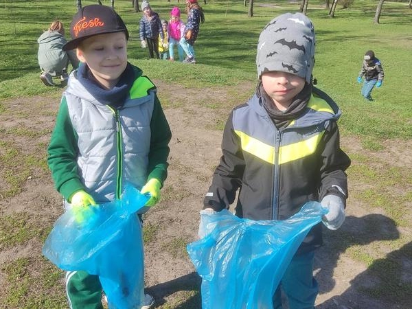 dwaj chłopcy w rekawicach trzymaja niebieskie worki na śmieci. W tle widać inne dzieci na trawiku.