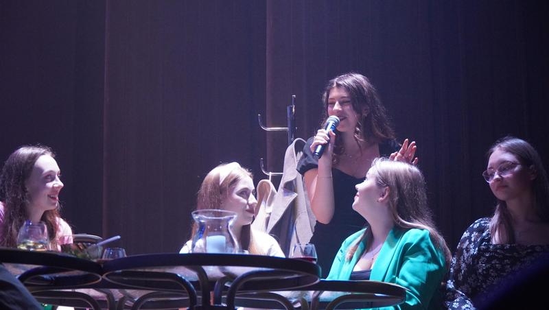 Dziewczęta siedzą przy stolikach. Sceneria jak w kawiarni.