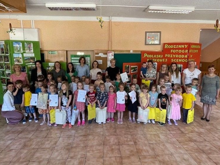 dzieci i opiekunki pozują do wspólnego zdjecia. Dzieci trzymaja dyplomy i torby z nagrodami. W tle widać wystawę fotogroafii