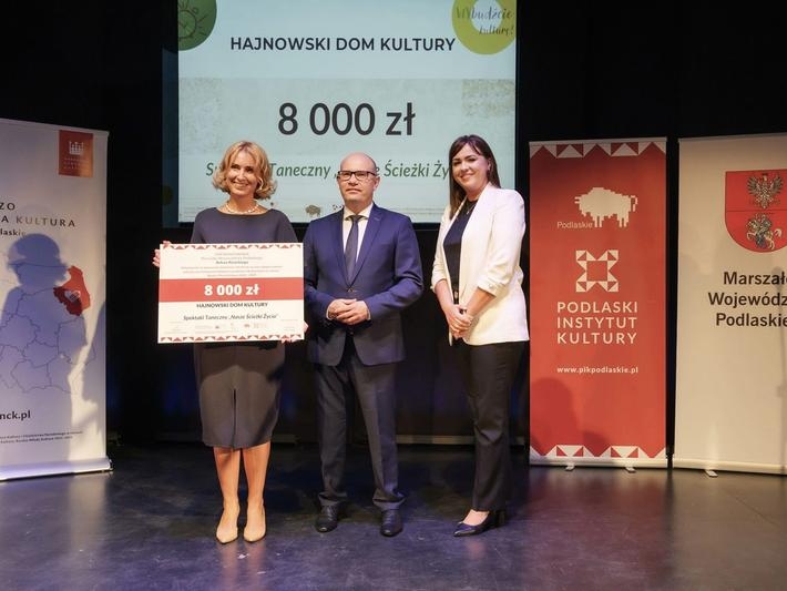 dwie kobieta i mężczyzna pozują do zdjecia na scenie. Kobieta po lewej trzyma czek z dotacją na 8 tys zł