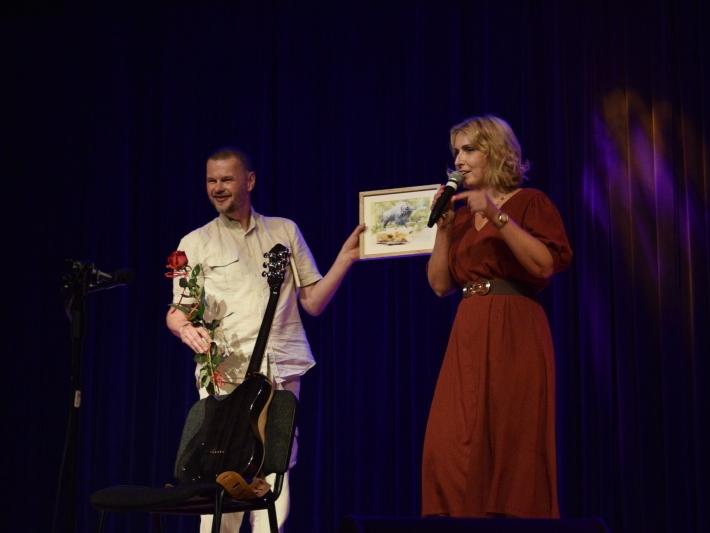 Na scenie Dyrektor Hajnowskiego Domu Kultury, Mariusz Oziu Orzechowski trzyma gitarę, różę, i obraz.