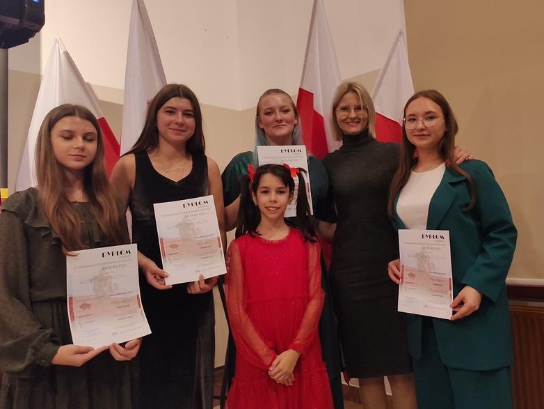 pięć dziewcząt z dyplomami pozuje do zdjęcia z instruktorką. W tle widać flagi narodowe.