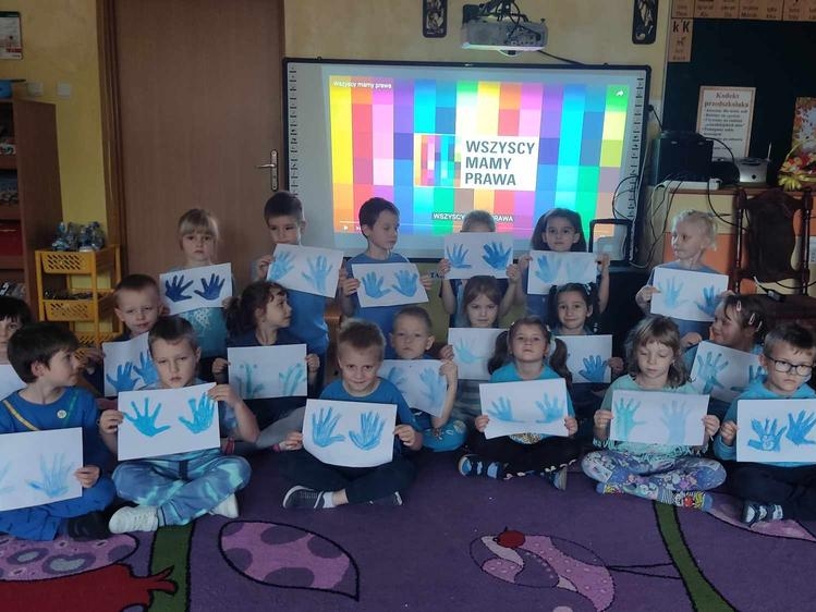 dzieci w niebieskich koszulkach trzymają w rękach kartki, na których są odciśniętymi na niebiesko dłońmi. W tle widać ekran z wyświetlanym slajdem na napiesem WSZYSCY MAMY PRAWA