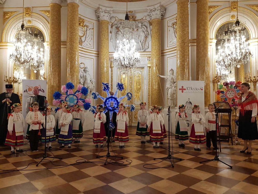 Dzieci z grupy „Kolebka” w odświętnych strojach ludowych stoją przed mikrofonami w Sali Wielkiej Zamku Królewskiego w Warszawie. Towarzyszą im nauczycielki, akordeonista i arcybiskup Sawa. W tle gwiazdy kolędnicze oraz bogato zdobione dekoracje ścienne, złote kolumny i posągi.