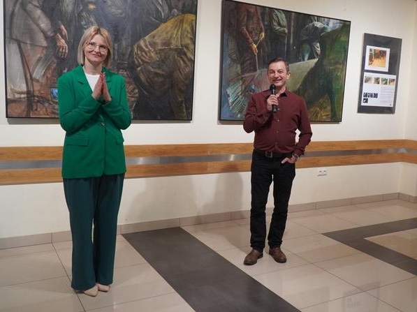 dwie osoby. Bije brawo, mężczyzna przemawia do migkkrofonu. w tle widać duże formaty obrazów