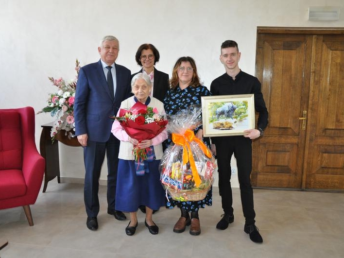 stulatka z bukietem kwiatów z rodziną i przedstawicielami Urzędu Miasta Hajnówka. Kobieta trzyma kosz ze słodyczami, młody meżczyzna trzyma obraz