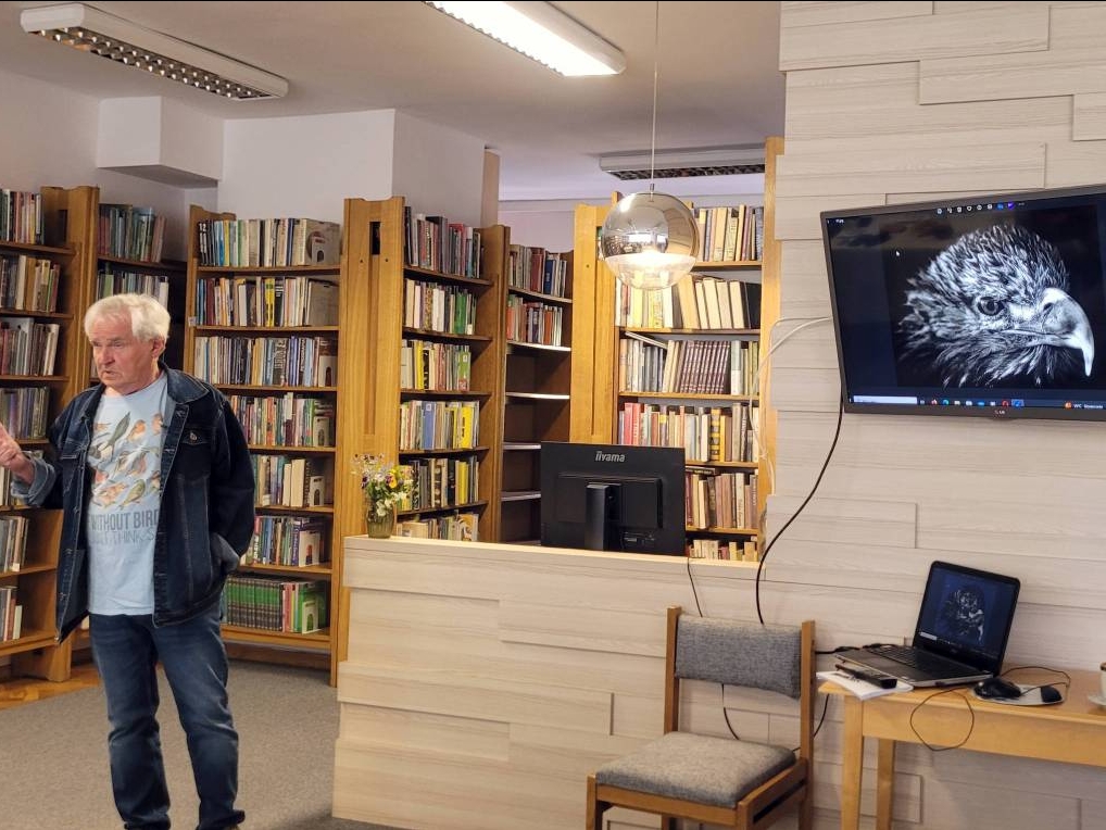 mężczyzna stoi w bobliotece. Na telewizorze wyświetlana jest głowa praka