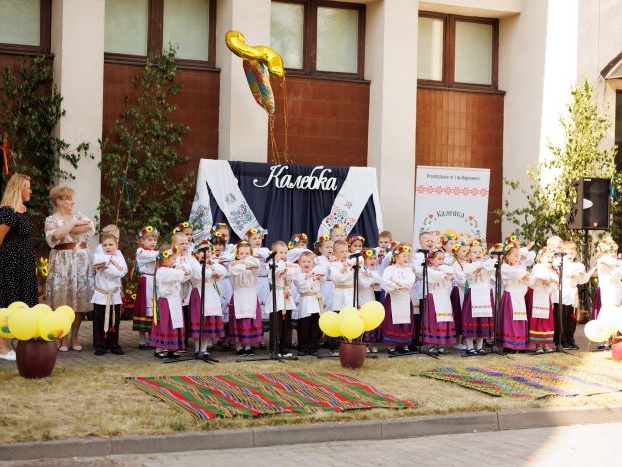 Dzieci z grupy Kolebka ubrane w ludowe stroje śpiewają przed mikrofonami, obok stoją dwie nauczycielki.