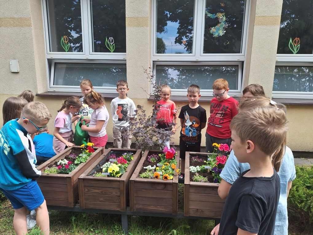 Grupa dzieci stoi wokół drewnianych donic z kwiatami. Dwie dziewczynki trzymają konewkę, którą podlewają kwiaty.