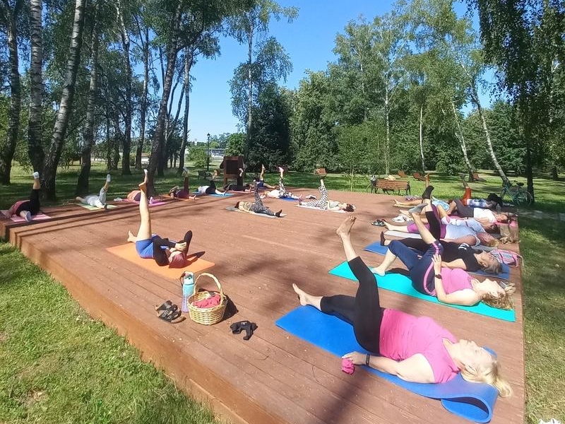 grupa osób leży na drewnianym podeście w parku miejskim. Wszyscy podnoszą nogi do góry
