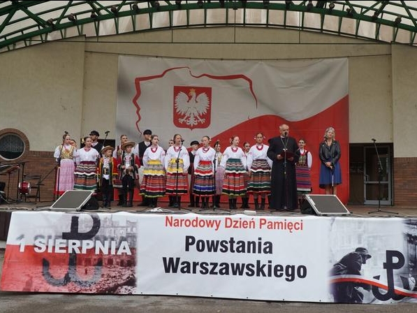 na scenie zespół, proboszcz i zapowiadająca koncert. Przed sceną zawieszony baner tematyczny dotyczący Powstania Warszawskiego. W tle widać wielką flagę z godłem Polski