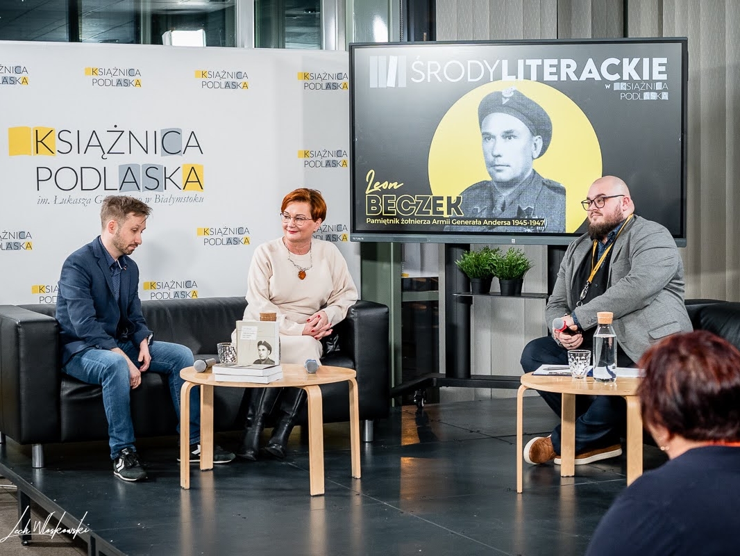trzy osoby siedzą przy stolikach. W tle wyświetlane jest zdjecie autora pamiątnika