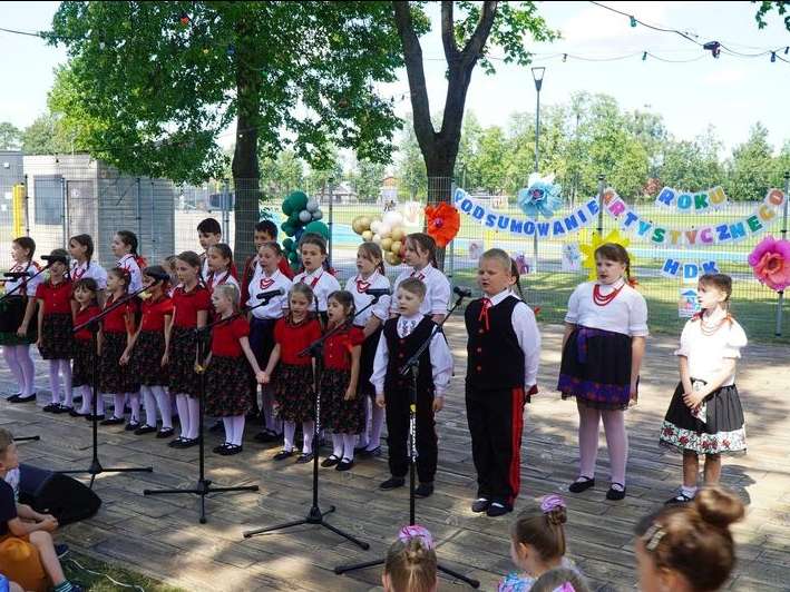 grupa dzieci w strojach estradowych występuje w plenerze. W tle widać napis: Podsumowanie roku artystycznego.