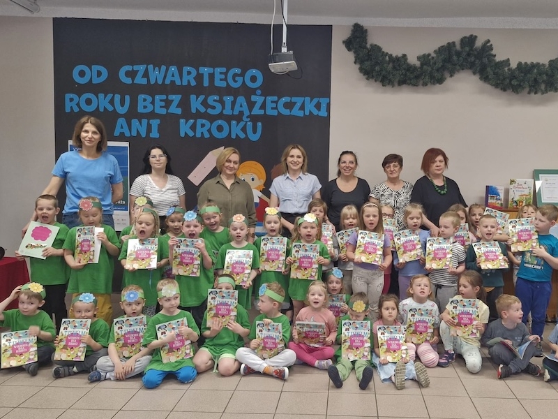 dzieci z dyplomami oraz opiekunki pozują do zdjecia W tle widać napis OD CZWARTEGO ROKU BEZ KSIĄŻECZEK ANI KROKU