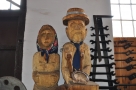 dwie drewniane rzeźby, przedstawiające sylwetkę kobiety w chustce oraz sylwetkę mężczyzny w kapeluszu oraz krawacie; stoją na tle białej ściany