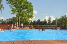 Na zdjęciu widać basen, znajduje sie w nim kilkoro dzieci. Na drugim planie widac cztery osoby, za nimi drzewa.
