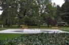 Na zdjęciu widac fontannę w parku, za nią stoja dwie ławki.