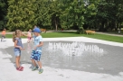 Na zdjęciu widac fontanne w parku. Po lewej stronie bawi się pięcioro dzieci.