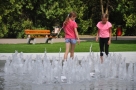 Na zdjęciu widac fontanne w parku. Obok niej chodzą dwie dziewczynki ubrane w różowe bluzki.
