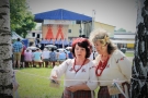 na pierwszym planie dwie kobiety w strojach ludowych, na szyi mają czerwone korale; w tle scena z niebieskim zadaszeniem, przed sceną na ławkach siedzą ludzie, wysłuchujący występujących artystów