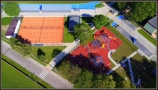 ozdjęcie z lotu ptaka: od lewej kort tenisowy pokryty podłożem w ceglastym kolorze; po prawej Rodzinna Sterefa Zabaw; dwa obiekty przedzielone są ogrodzeniem z siatki; na wysokości placu zabaw ulokowane są miejsca parkingowe