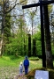 Chłopiec i dziewczynka znajdujący się w miejscu pamięci straceń mieszkańców Hajnówki i okolic; obok stoi wysoki drewniany krzyż z tablicą pamiątkową, wokół zieleń oraz drzewa