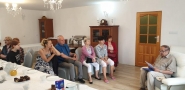 grupa uczestników zebrana w sali, siedzą na krzesłach, przed nimi na fotelu siedzi mężczyzna czytający książkę; z lewej strony stoi stół, na którym jest poczęstunek